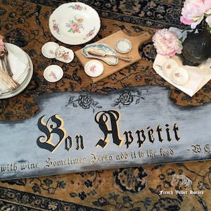 May include: A gray wooden sign with the words "Bon Appetit" in black script. The sign also includes the text "I cook with wine. Sometimes I even add it to the food - W.C. Fields".