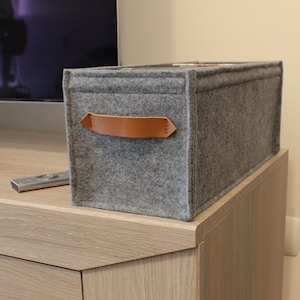 May include: A rectangular gray felt storage bin with a brown leather handle. The bin is sitting on a light wood cabinet. A TV and remote control are in the background. The bin is designed for organizing items.