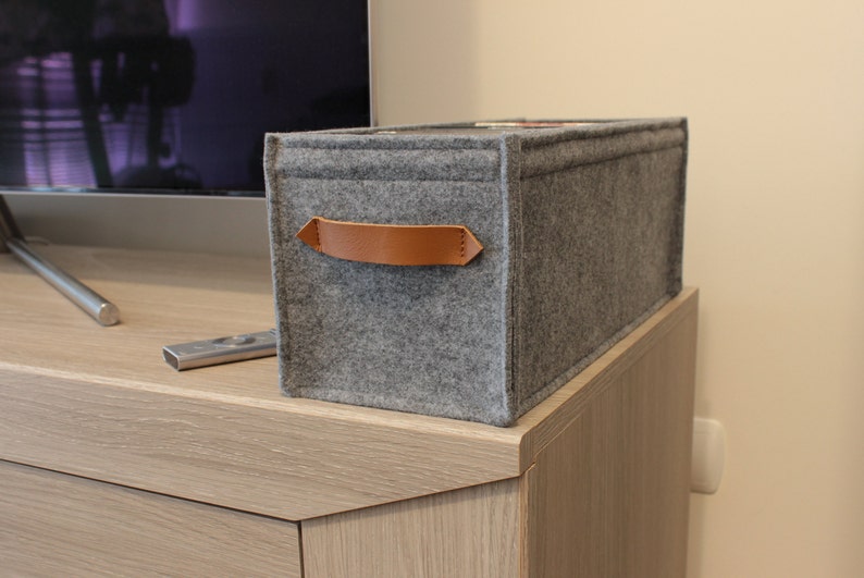 May include: A rectangular gray felt storage bin with a brown leather handle. The bin is sitting on a light wood cabinet. A TV and remote control are in the background. The bin is designed for organizing items.