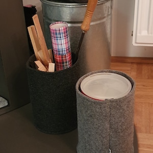 Puede incluir: Dos contenedores de almacenamiento de fieltro gris, uno con un rollo de papel de regalo a cuadros y palitos de madera en su interior, el otro con una tapa blanca. Los contenedores están sobre una superficie marrón.