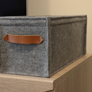 May include: A rectangular gray felt storage bin with a brown leather handle. The bin is sitting on a light-colored wooden surface. The handle is attached to the front of the bin.