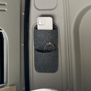 May include: A gray felt organizer with two pockets mounted on a light gray wall. A white smartphone is in the top pocket, and a pair of eyeglasses is in the lower pocket. The organizer is designed for storage and organization.