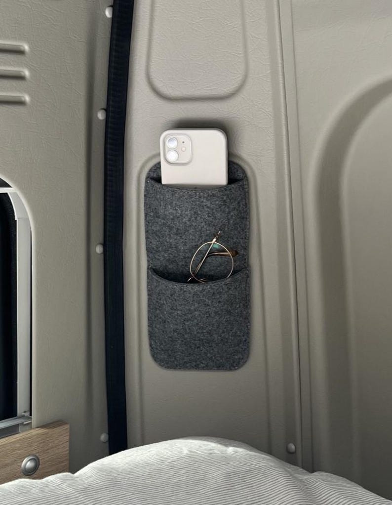 May include: A gray felt organizer with two pockets mounted on a light gray wall. A white smartphone is in the top pocket, and a pair of eyeglasses is in the lower pocket. The organizer is designed for storage and organization.