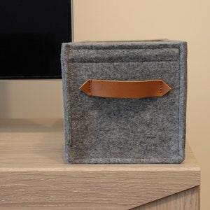 May include: A gray felt storage cube with a brown leather handle. The cube is sitting on a light wood surface. The handle is attached with stitching. The cube is a simple, modern design.