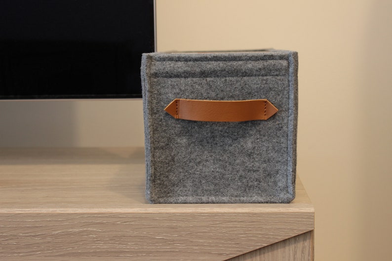 May include: A gray felt storage cube with a brown leather handle. The cube is sitting on a light wood surface. The handle is attached with stitching. The cube is a simple, modern design.