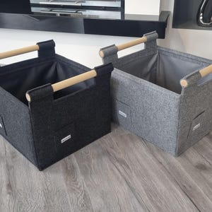 May include: Two rectangular storage bins, one dark gray and one light gray, with wooden handles. The bins are made of a felt-like material and have a small pocket on the front. The handles are made of light-colored wood.