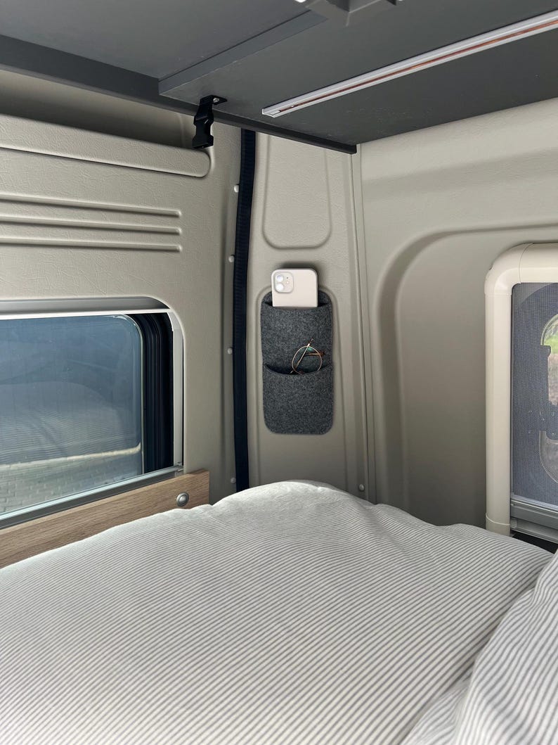 May include: Interior view of a camper van with a gray felt pocket organizer mounted on the wall. A smartphone and eyeglasses are in the pockets. A bed with a striped comforter is in the foreground. A window is visible.