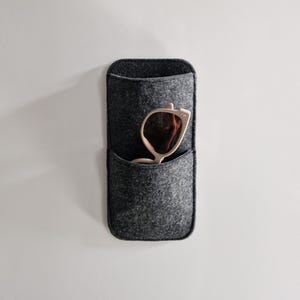 May include: A gray felt sunglasses holder with a pocket, mounted on a white wall. A pair of light-colored sunglasses with brown lenses is partially visible inside the pocket. The holder has a rectangular shape with rounded edges.