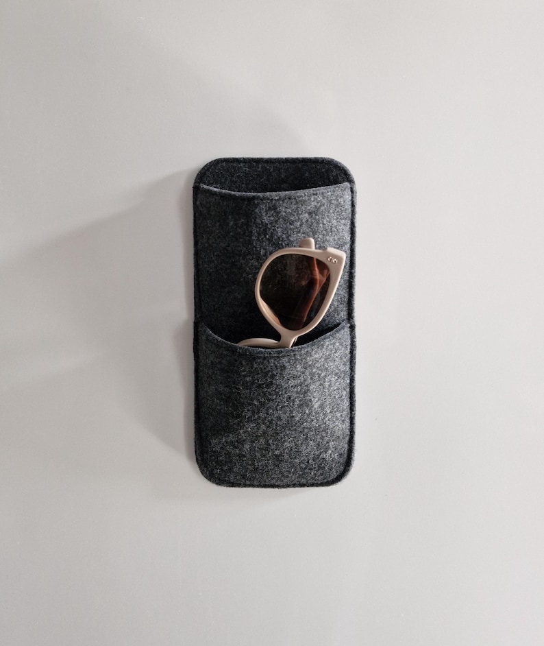 May include: A gray felt sunglasses holder with a pocket, mounted on a white wall. A pair of light-colored sunglasses with brown lenses is partially visible inside the pocket. The holder has a rectangular shape with rounded edges.