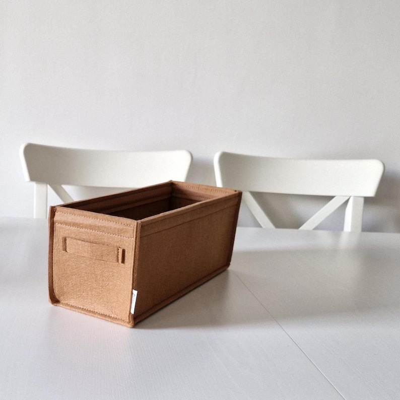 May include: A rectangular, brown felt organizer with a handle on each side. The organizer is sitting on a white surface. Two white chairs are in the background.