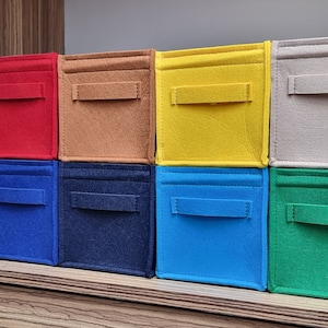 May include: A collection of ten colorful felt storage bins in various shades, including red, black, blue, and green. Each bin has a rectangular shape with a handle on the front. The bins are stacked on a wooden shelf.