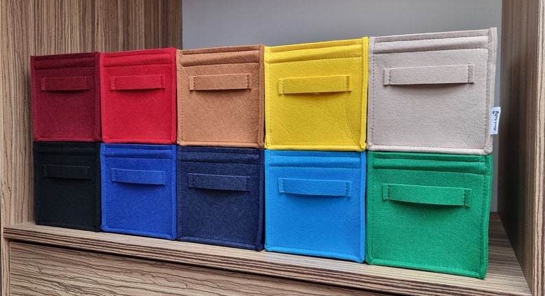 May include: A collection of ten colorful felt storage bins in various shades, including red, black, blue, and green. Each bin has a rectangular shape with a handle on the front. The bins are stacked on a wooden shelf.