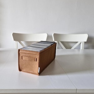May include: A rectangular, tan felt CD storage box with a handle on the side, filled with CDs. The box is on a white table, with two white chairs in the background. The box is approximately 12 inches long.