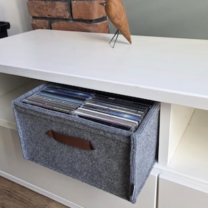 May include: A gray felt storage bin with a brown leather handle, filled with CDs, sits in a white shelf unit. A wooden bird sculpture with metal legs rests on the shelf above. A brick wall is in the background.