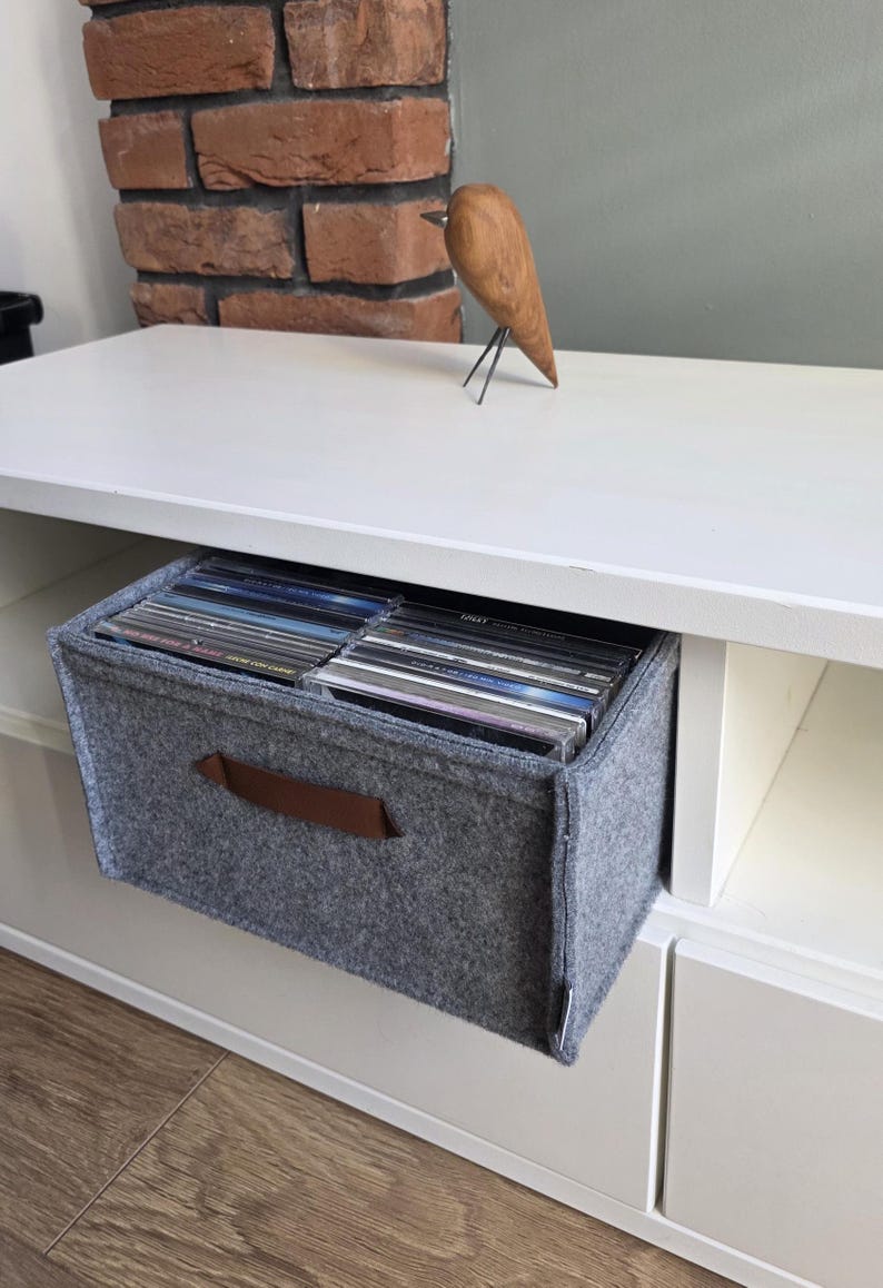 May include: A gray felt storage bin with a brown leather handle, filled with CDs, sits in a white shelf unit. A wooden bird sculpture with metal legs rests on the shelf above. A brick wall is in the background.
