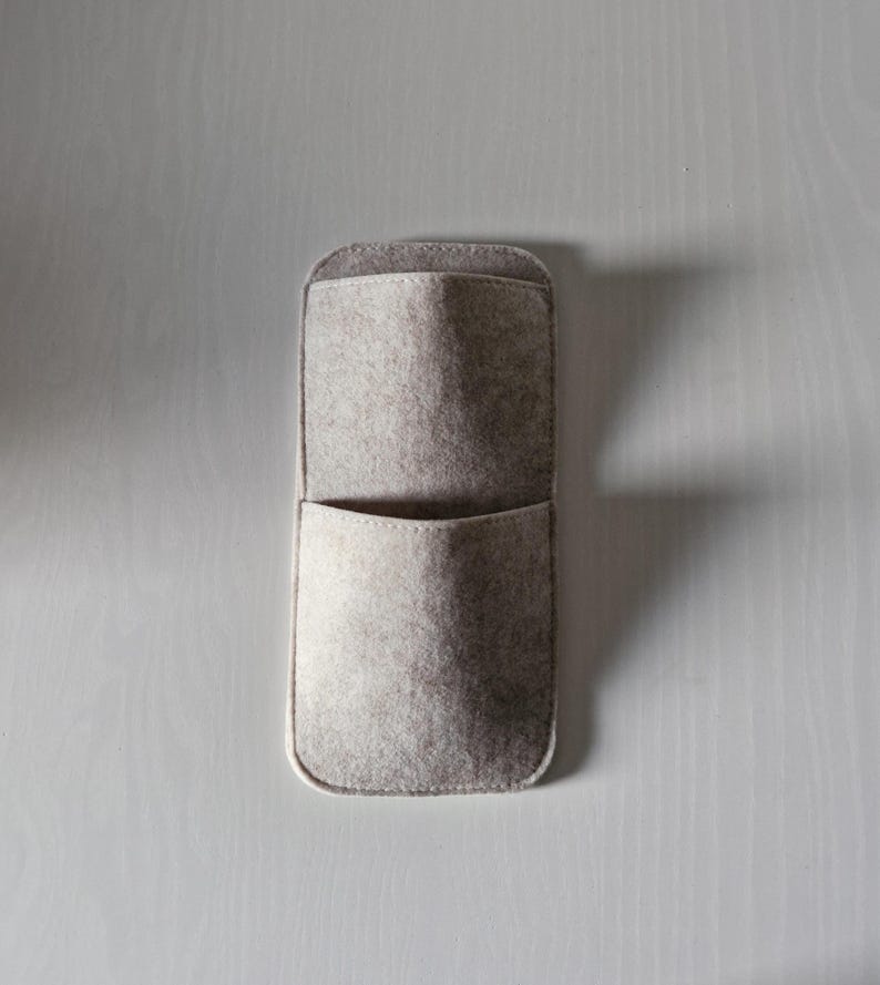 May include: A rectangular, light gray felt wall organizer with two pockets. The top pocket is larger and open, while the bottom pocket is smaller. The organizer is made of soft felt material and has a rounded top and bottom.