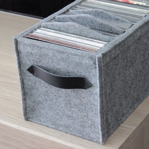 Peut inclure: Un coffret de rangement pour CD en feutre gris avec une poignée en cuir noir. La boîte est remplie de CD et comporte des séparateurs. La boîte est posée sur une surface en bois clair.