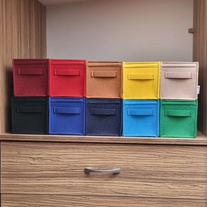 May include: A collection of ten colorful felt storage bins in various shades, including red, black, blue, and yellow. Each bin has a rectangular shape with a small handle on the front. The bins are arranged on a wooden shelf.