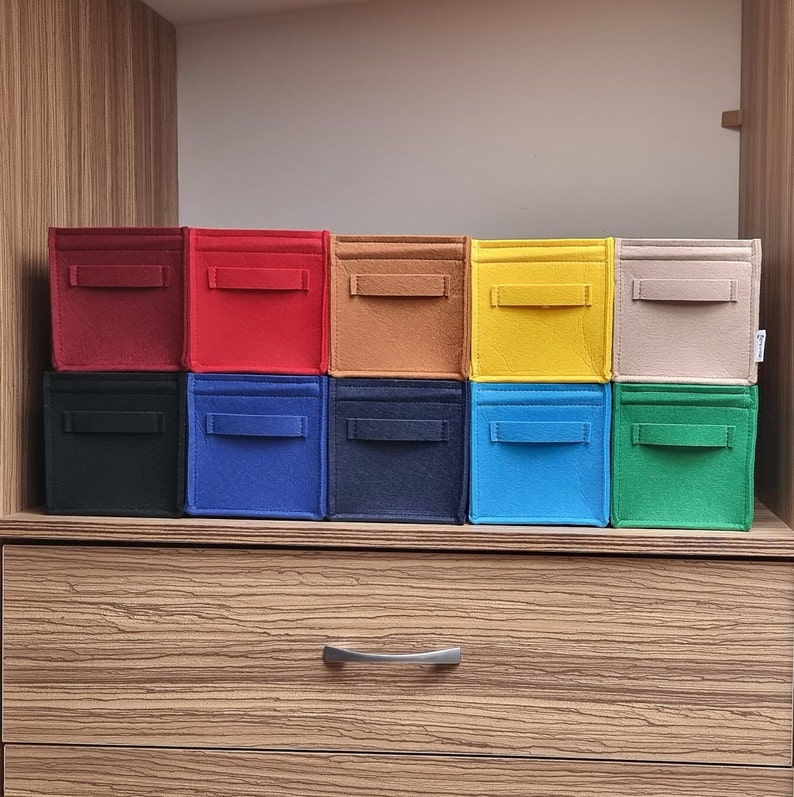 May include: A collection of ten colorful felt storage bins in various shades, including red, black, blue, and yellow. Each bin has a rectangular shape with a small handle on the front. The bins are arranged on a wooden shelf.