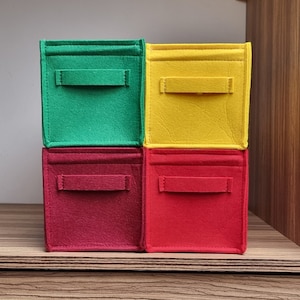 May include: Four felt storage boxes stacked on top of each other. The top box is yellow, the second box is green, the third box is red, and the bottom box is red. Each box has a handle.