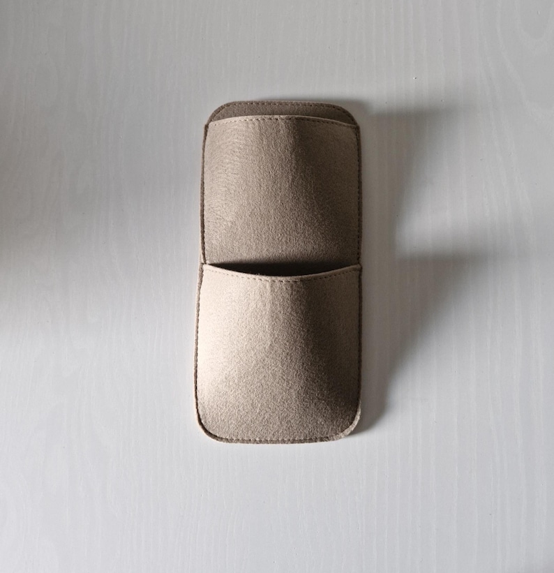May include: A beige felt wall organizer with two pockets. The top pocket is open, and the bottom pocket is slightly smaller. The organizer is rectangular with rounded corners and is designed to be mounted on a wall.