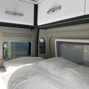 May include: Interior view of a camper van with a bed, pillows, and overhead storage cabinets. The bed is covered with a striped comforter and pillows. Windows are visible, offering a view of the outside. The cabinets are white with silver handles.