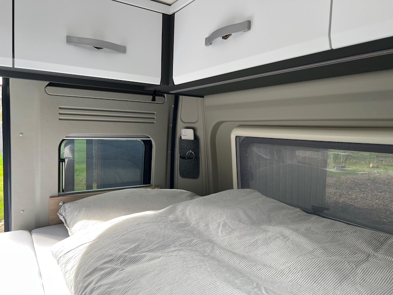 May include: Interior view of a camper van with a bed, pillows, and overhead storage cabinets. The bed is covered with a striped comforter and pillows. Windows are visible, offering a view of the outside. The cabinets are white with silver handles.