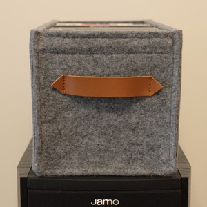 May include: A gray felt storage box with a brown leather handle. The box is rectangular and sits atop a black speaker. The handle is stitched to the felt. The box appears to be holding records.