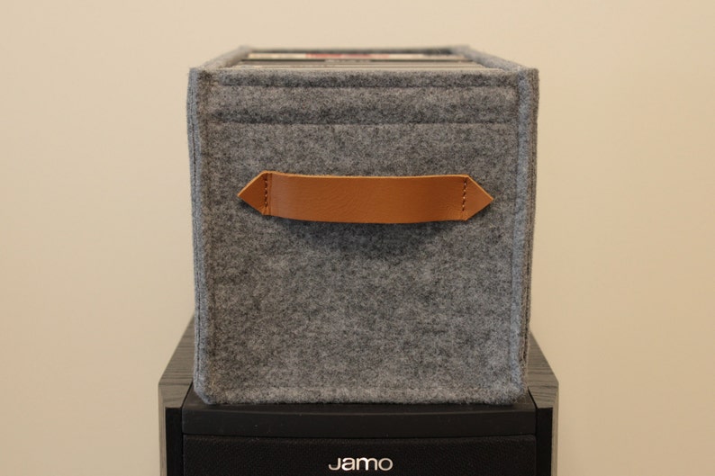 May include: A gray felt storage box with a brown leather handle. The box is rectangular and sits atop a black speaker. The handle is stitched to the felt. The box appears to be holding records.