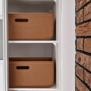 Könnte beinhalten: Zwei rechteckige, braune Aufbewahrungsboxen mit abgerundeten Ecken und einem Griffausschnitt sind auf weißen Regalen gestapelt. Die Boxen bestehen aus einem filzartigen Material. Der Hintergrund umfasst eine Backsteinmauer und einen weißen Schrank.