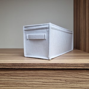 May include: A rectangular, light gray felt storage bin with a handle on the side. The bin is placed on a wooden shelf. The bin is designed to organize items and is a neutral color.