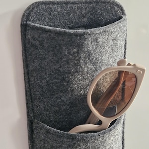 May include: A gray felt wall organizer with a pocket, holding a pair of beige sunglasses. The sunglasses have a light-colored frame and brown lenses. The organizer is designed to hold glasses or other small items.
