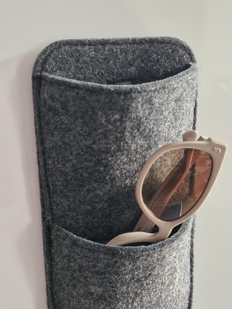 May include: A gray felt wall organizer with a pocket, holding a pair of beige sunglasses. The sunglasses have a light-colored frame and brown lenses. The organizer is designed to hold glasses or other small items.