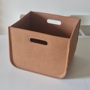 Könnte beinhalten: Rechteckige Aufbewahrungsbox aus hellbraunem Filz mit zwei ovalen Griffen. Die Box hat abgerundete Ecken und sichtbare Nähte. Sie steht auf einer weißen Oberfläche. Maße in Zentimetern.