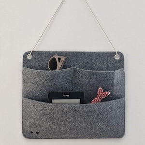 Pode incluir: Um organizador de parede retangular de feltro cinza com duas fileiras de bolsos, ilhós de metal e um cordão para pendurar. Os bolsos contêm óculos de sol claros, um leitor de e-book Kindle preto e um pequeno peixe de tecido vermelho estampado.