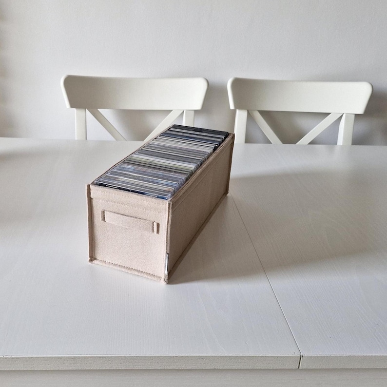 May include: A beige felt CD storage box filled with CDs, placed on a white table. The box has a rectangular shape with two small handle cutouts on the sides. Two white chairs are in the background.