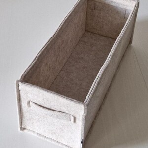 May include: A rectangular, light gray felt storage bin with a handle on one side. The bin is open at the top and has a simple, minimalist design. The felt material appears soft and the bin is empty.