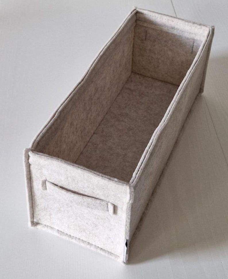 May include: A rectangular, light gray felt storage bin with a handle on one side. The bin is open at the top and has a simple, minimalist design. The felt material appears soft and the bin is empty.
