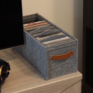 May include: A gray felt record storage box with a brown leather handle. The box is filled with vinyl records and sits on a light wood surface. A pair of black headphones with gold accents are in the foreground.