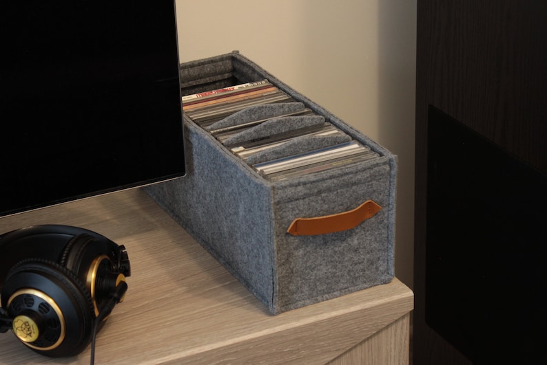 May include: A gray felt record storage box with a brown leather handle. The box is filled with vinyl records and sits on a light wood surface. A pair of black headphones with gold accents are in the foreground.