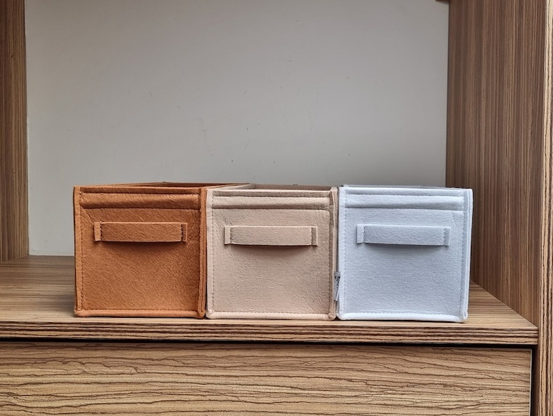May include: Three felt storage cubes in shades of brown, beige, and light blue. Each cube has a rectangular handle on the front. The cubes are arranged on a wooden shelf, offering a simple and organized storage solution.