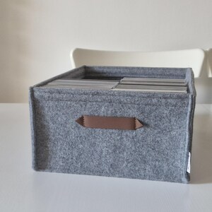 May include: A gray felt storage box with a brown handle. The box is filled with cards or papers. The box is sitting on a white surface. The box is approximately 12 inches wide, 10 inches deep, and 8 inches tall.