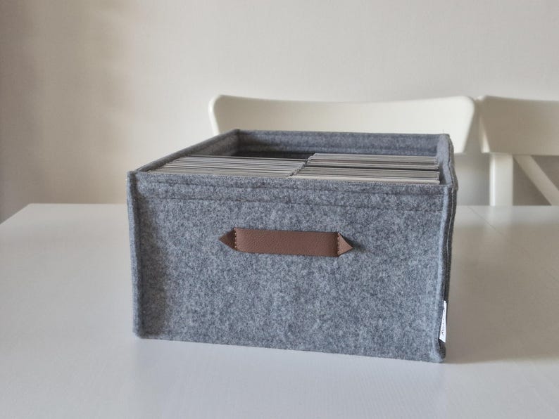 May include: A gray felt storage box with a brown handle. The box is filled with cards or papers. The box is sitting on a white surface. The box is approximately 12 inches wide, 10 inches deep, and 8 inches tall.