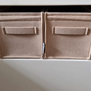 May include: Two beige felt storage bins with rectangular handles are placed side by side. The bins are inside a white cabinet. A small tag with text is visible on the right bin. The bins are designed to organize items.