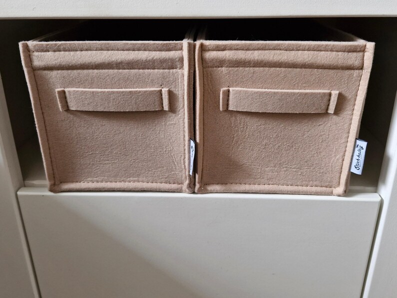 May include: Two beige felt storage bins with rectangular handles are placed side by side. The bins are inside a white cabinet. A small tag with text is visible on the right bin. The bins are designed to organize items.