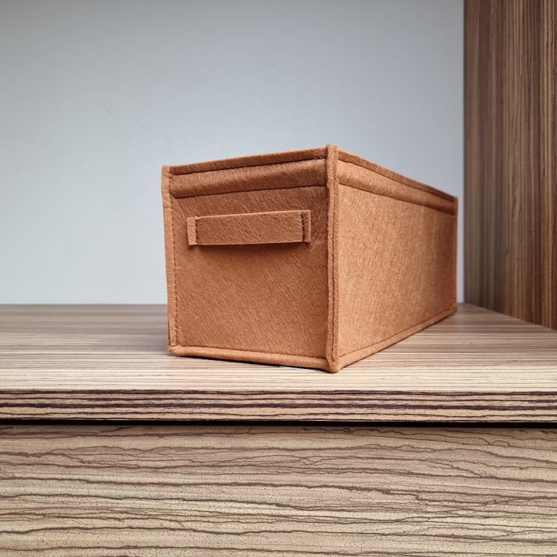 May include: A rectangular, brown felt storage organizer with a handle on one side. The organizer is sitting on a wooden shelf with a striped pattern. The background is a light gray wall and a wooden cabinet.