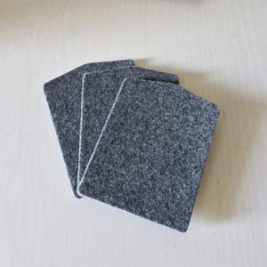 May include: Three gray felt coasters arranged on a light wood surface. The coasters are rectangular with rounded corners and a textured appearance. They are a solid, neutral color, suitable for various home decor styles.