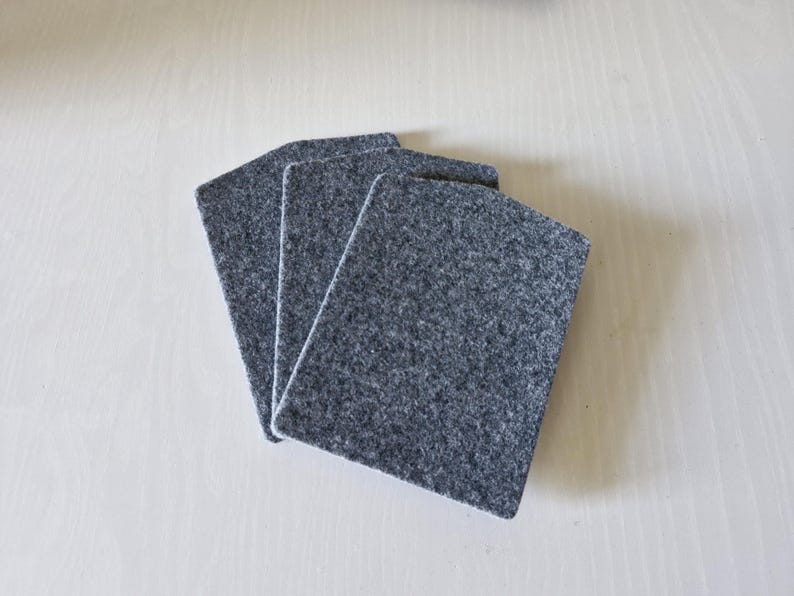 May include: Three gray felt coasters arranged on a light wood surface. The coasters are rectangular with rounded corners and a textured appearance. They are a solid, neutral color, suitable for various home decor styles.