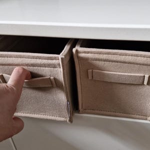 May include: Two beige fabric storage bins with handles are placed inside a white cabinet. A hand with a silver bracelet is reaching for one of the bins. The bins are rectangular and have a simple, minimalist design.