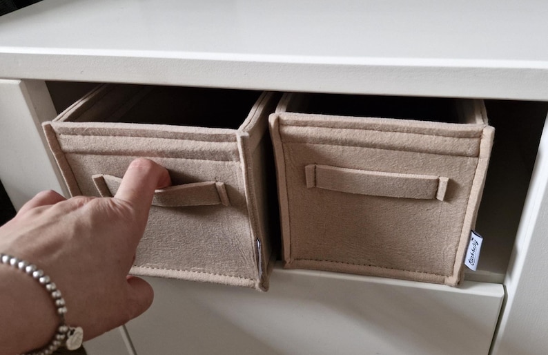 May include: Two beige fabric storage bins with handles are placed inside a white cabinet. A hand with a silver bracelet is reaching for one of the bins. The bins are rectangular and have a simple, minimalist design.